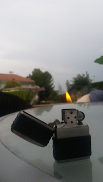 A metallic lighter with a black grip is lit on top of a glass table. The background features a blurred outdoor setting with trees, a house with a red roof, and a cloudy sky.