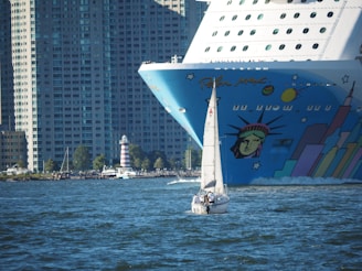 A large cruise ship with vibrant artwork on its bow, featuring a stylized version of the Statue of Liberty, is juxtaposed with a small sailing boat in the foreground. The background is dominated by tall, densely packed urban buildings, highlighting a mix of urban life and maritime travel.