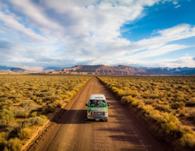 A lone vehicle travels down a straight dirt road surrounded by an expansive desert landscape, with rugged mountain ranges in the background under a partly cloudy sky. The scene conveys a sense of freedom and adventure.