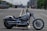 A Harley-Davidson motorcycle is parked on an empty road with city buildings in the background. The scene includes other vehicles such as trucks in the distance. The motorcycle has a sleek, polished chrome finish and large wheels.