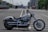 A Harley-Davidson motorcycle is parked on an empty road with city buildings in the background. The scene includes other vehicles such as trucks in the distance. The motorcycle has a sleek, polished chrome finish and large wheels.