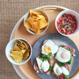 A wooden tray holds a plate with toast topped with eggs, prosciutto, spinach, and cheese. Surrounding the plate are bowls filled with tortilla chips and a tomato salsa.