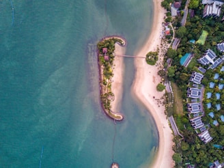 An aerial view of a coastal area with a small island connected by a footbridge to the mainland. The island is lush with vegetation and has a couple of small structures. The surrounding waters are a deep blue-green, contrasted by the sandy beaches of the mainland. Residential buildings and greenery are visible in the background.