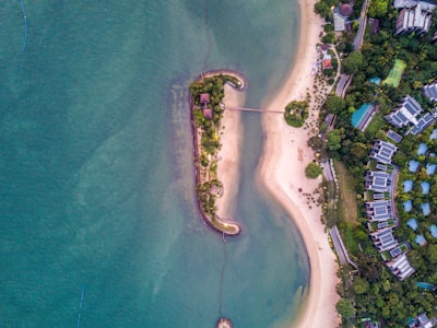 An aerial view of a coastal area with a small island connected by a footbridge to the mainland. The island is lush with vegetation and has a couple of small structures. The surrounding waters are a deep blue-green, contrasted by the sandy beaches of the mainland. Residential buildings and greenery are visible in the background.