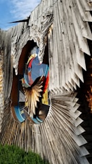 An abstract wooden sculpture with intricate, layered designs creating a three-dimensional effect. The sculpture incorporates vibrant stained glass elements with bold colors, and has a large, circular cut-out in the center. The background features a clear blue sky and some greenery at the base.