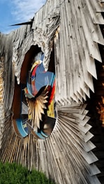 An abstract wooden sculpture with intricate, layered designs creating a three-dimensional effect. The sculpture incorporates vibrant stained glass elements with bold colors, and has a large, circular cut-out in the center. The background features a clear blue sky and some greenery at the base.