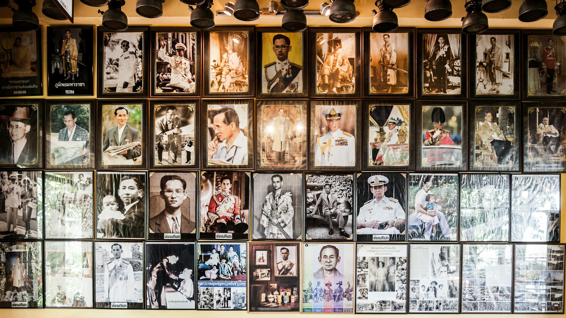 A collection of framed photographs on a wall, featuring a variety of historical and formal portraits of a person in different settings, including military uniforms and traditional attire. Overhead lighting fixtures are visible at the top of the image.