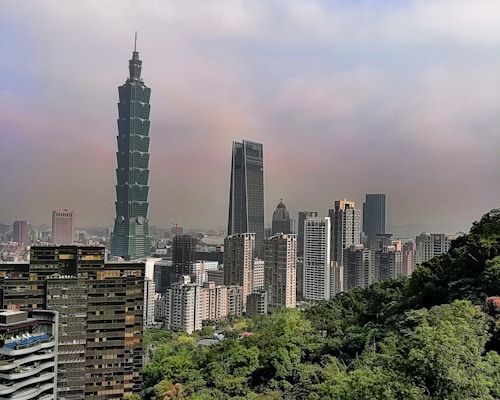 A cityscape featuring a prominent skyscraper with a unique tiered design amidst several other modern high-rise buildings. The scene includes lush greenery in the foreground, with hazy skies above the city.