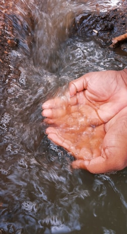 Two cupped hands are immersed in a flowing stream, catching fresh water. The hands are positioned against the backdrop of moving water, with the surrounding environment being earthy and natural.