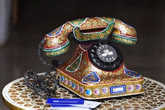 A vintage rotary dial telephone adorned with intricate, colorful patterns sits on an equally ornate table. The phone features a prominent black dial with numbers and is embellished with floral and geometric designs in vibrant shades of red, green, gold, and blue.
