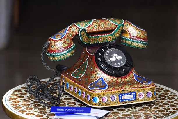 A vintage rotary dial telephone adorned with intricate, colorful patterns sits on an equally ornate table. The phone features a prominent black dial with numbers and is embellished with floral and geometric designs in vibrant shades of red, green, gold, and blue.