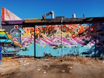 A colorful graffiti mural covers the entire side of a building. The artwork includes vibrant shades of blue, orange, pink, and purple, with intricate lettering and designs. The graffiti features a cartoonish spray paint can with expressive eyes, adding a playful element to the mural. The area in front of the wall is scattered with debris and has a gritty urban feel.