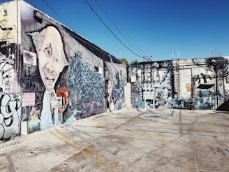 A vibrant mural covers the walls of an urban alleyway, featuring surreal and abstract elements. A large, exaggerated face with expressive eyes and a mustache is prominently displayed alongside intricate graffiti art. The scene includes intricate mechanical designs and figures with detailed expressions.