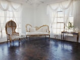 An elegant room featuring vintage furniture with ornate gold detailing, sheer white curtains draping from large windows, and a dark polished floor. A plush white settee and a similarly styled high-backed chair create a refined and luxurious ambiance. A small side table and a decorative console table complete the setting, adorned with delicate decor pieces.
