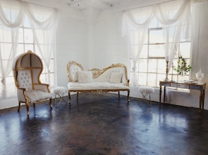 An elegant room featuring vintage furniture with ornate gold detailing, sheer white curtains draping from large windows, and a dark polished floor. A plush white settee and a similarly styled high-backed chair create a refined and luxurious ambiance. A small side table and a decorative console table complete the setting, adorned with delicate decor pieces.