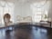 An elegant room featuring vintage furniture with ornate gold detailing, sheer white curtains draping from large windows, and a dark polished floor. A plush white settee and a similarly styled high-backed chair create a refined and luxurious ambiance. A small side table and a decorative console table complete the setting, adorned with delicate decor pieces.
