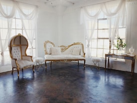 An elegant room featuring vintage furniture with ornate gold detailing, sheer white curtains draping from large windows, and a dark polished floor. A plush white settee and a similarly styled high-backed chair create a refined and luxurious ambiance. A small side table and a decorative console table complete the setting, adorned with delicate decor pieces.