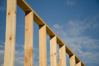 Wooden beams form an incomplete structure against a backdrop of a blue sky with scattered clouds. The beams angle diagonally across the image, highlighting their texture and natural wood grain.