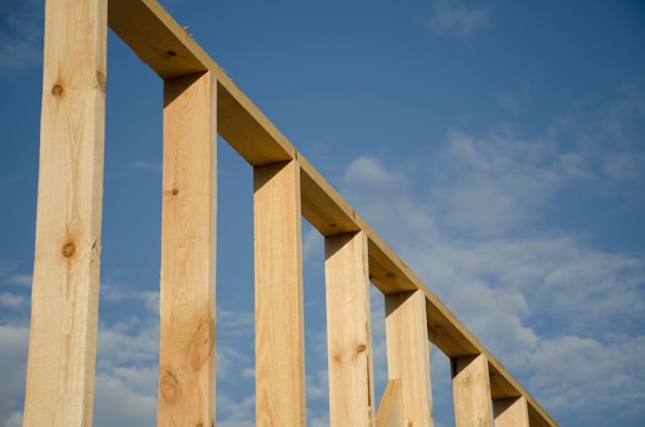 Wooden beams form an incomplete structure against a backdrop of a blue sky with scattered clouds. The beams angle diagonally across the image, highlighting their texture and natural wood grain.