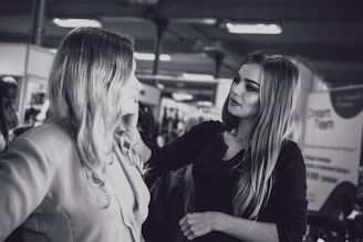 A makeup artist is applying cosmetics to a woman with long hair. They are in an indoor setting with blurred background elements, possibly a salon or a professional event. The photo is in black and white.