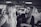 A makeup artist is applying cosmetics to a woman with long hair. They are in an indoor setting with blurred background elements, possibly a salon or a professional event. The photo is in black and white.
