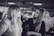 A makeup artist is applying cosmetics to a woman with long hair. They are in an indoor setting with blurred background elements, possibly a salon or a professional event. The photo is in black and white.