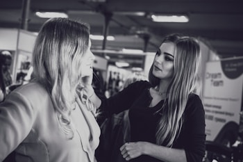 A makeup artist is applying cosmetics to a woman with long hair. They are in an indoor setting with blurred background elements, possibly a salon or a professional event. The photo is in black and white.