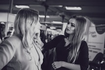 A makeup artist is applying cosmetics to a woman with long hair. They are in an indoor setting with blurred background elements, possibly a salon or a professional event. The photo is in black and white.