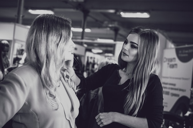 A makeup artist is applying cosmetics to a woman with long hair. They are in an indoor setting with blurred background elements, possibly a salon or a professional event. The photo is in black and white.
