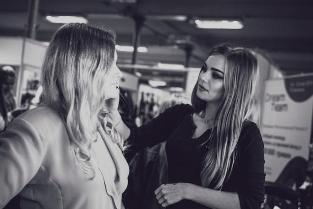 A makeup artist is applying cosmetics to a woman with long hair. They are in an indoor setting with blurred background elements, possibly a salon or a professional event. The photo is in black and white.