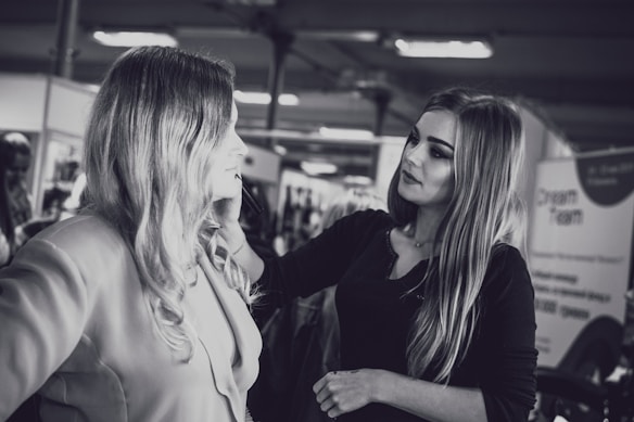 A makeup artist is applying cosmetics to a woman with long hair. They are in an indoor setting with blurred background elements, possibly a salon or a professional event. The photo is in black and white.