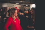 A woman is applying makeup to another woman in a well-lit indoor setting, likely at a beauty or fashion event. The woman receiving the makeup is wearing a bright red outfit, while the surroundings have several other people and promotional posters or banners.
