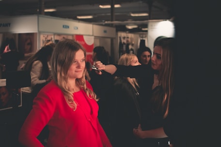 A woman is applying makeup to another woman in a well-lit indoor setting, likely at a beauty or fashion event. The woman receiving the makeup is wearing a bright red outfit, while the surroundings have several other people and promotional posters or banners.