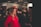 A woman is applying makeup to another woman in a well-lit indoor setting, likely at a beauty or fashion event. The woman receiving the makeup is wearing a bright red outfit, while the surroundings have several other people and promotional posters or banners.