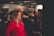 A woman is applying makeup to another woman in a well-lit indoor setting, likely at a beauty or fashion event. The woman receiving the makeup is wearing a bright red outfit, while the surroundings have several other people and promotional posters or banners.