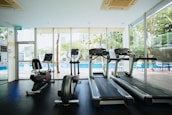 A bright and modern gym space featuring three treadmills and an elliptical machine, positioned in front of large glass windows that reveal a view of a pool and outdoor seating. The interior is well-lit with natural light, enhancing the clean and organized ambiance.