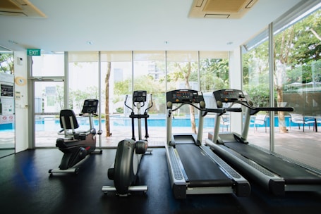 A bright and modern gym space featuring three treadmills and an elliptical machine, positioned in front of large glass windows that reveal a view of a pool and outdoor seating. The interior is well-lit with natural light, enhancing the clean and organized ambiance.