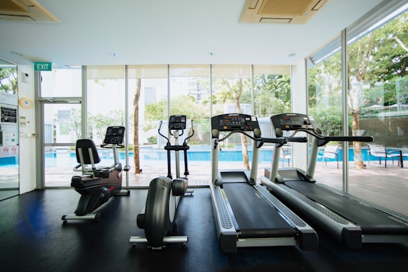 A bright and modern gym space featuring three treadmills and an elliptical machine, positioned in front of large glass windows that reveal a view of a pool and outdoor seating. The interior is well-lit with natural light, enhancing the clean and organized ambiance.
