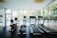 A bright and modern gym space featuring three treadmills and an elliptical machine, positioned in front of large glass windows that reveal a view of a pool and outdoor seating. The interior is well-lit with natural light, enhancing the clean and organized ambiance.