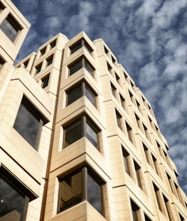 A towering, modern office building with a beige facade capped with large, symmetrical windows, set against a backdrop of a partly cloudy blue sky.