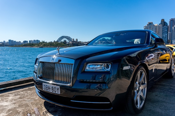 A luxury car is parked by a waterfront with a view of a city skyline and a prominent bridge in the background. The sky is clear and blue, casting reflections on the polished surface of the car.