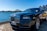 A luxury car is parked by a waterfront with a view of a city skyline and a prominent bridge in the background. The sky is clear and blue, casting reflections on the polished surface of the car.