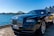 A luxury car is parked by a waterfront with a view of a city skyline and a prominent bridge in the background. The sky is clear and blue, casting reflections on the polished surface of the car.