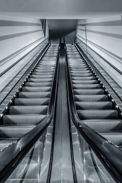 A modern and sleek escalator with metallic finishes ascends upwards. The setting appears to be indoors, with the escalator positioned symmetrically in the frame. The architecture is minimalistic with strong lines and a polished look.