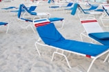 Multiple beach loungers with white frames and blue fabric covers are arranged on sandy beach. Some have red accents, and the area appears unoccupied with distant ocean visible.