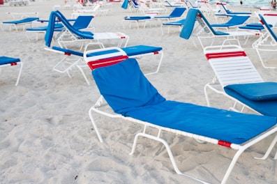 Multiple beach loungers with white frames and blue fabric covers are arranged on sandy beach. Some have red accents, and the area appears unoccupied with distant ocean visible.
