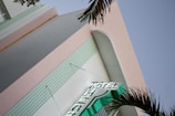 A building with a pastel pink facade and vertical elements outlined with horizontal grooves. A neon sign displaying the word 'HOTEL' protrudes from the building, and palm fronds are visible in the foreground, indicating a likely tropical or seaside location.