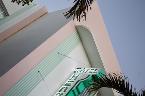 A building with a pastel pink facade and vertical elements outlined with horizontal grooves. A neon sign displaying the word 'HOTEL' protrudes from the building, and palm fronds are visible in the foreground, indicating a likely tropical or seaside location.