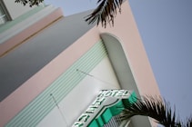 A building with a pastel pink facade and vertical elements outlined with horizontal grooves. A neon sign displaying the word 'HOTEL' protrudes from the building, and palm fronds are visible in the foreground, indicating a likely tropical or seaside location.