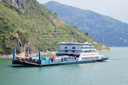 A large ferry boat is carrying several trucks across a wide river. The boat has a multi-story structure and is painted in shades of blue and white. There are green, hilly terrains on the left side and mountainous landscapes in the background with a calm sky.